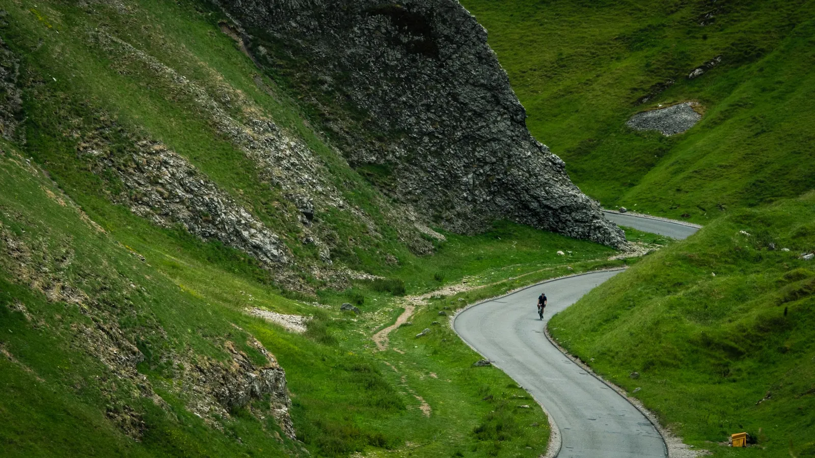 Fotografía ambiental de un ciclista de carretera rodando en solitario por una carretera de montaña que serpentea entre laderas verdes
