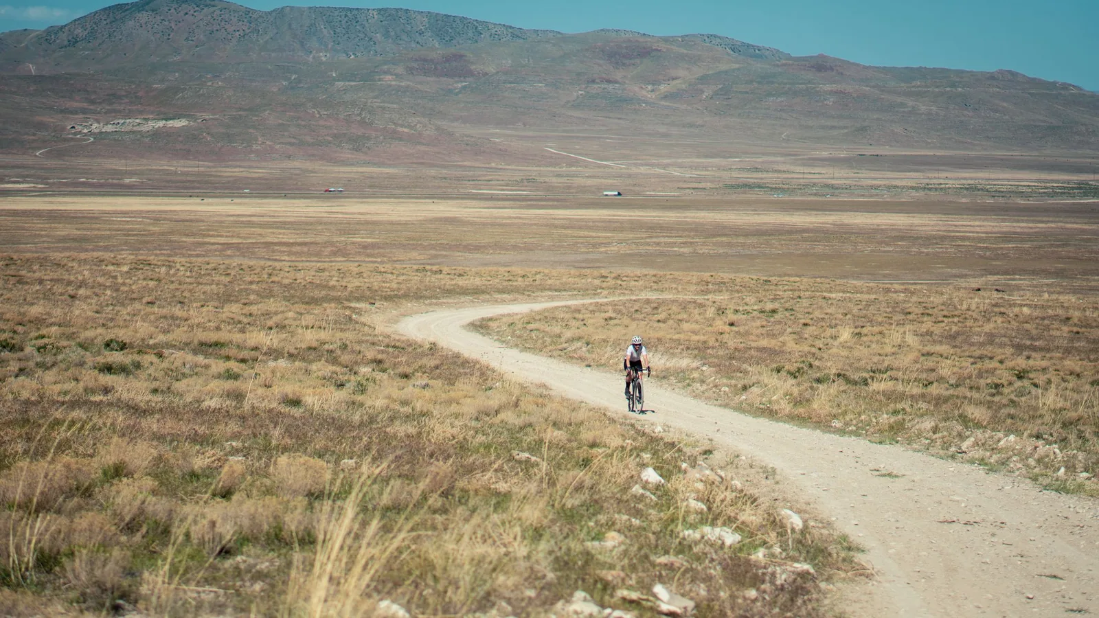 Fotografía ambiental de un ciclista avanzando por una pista de grava que cruza una llanura seca con una sierra al fondo