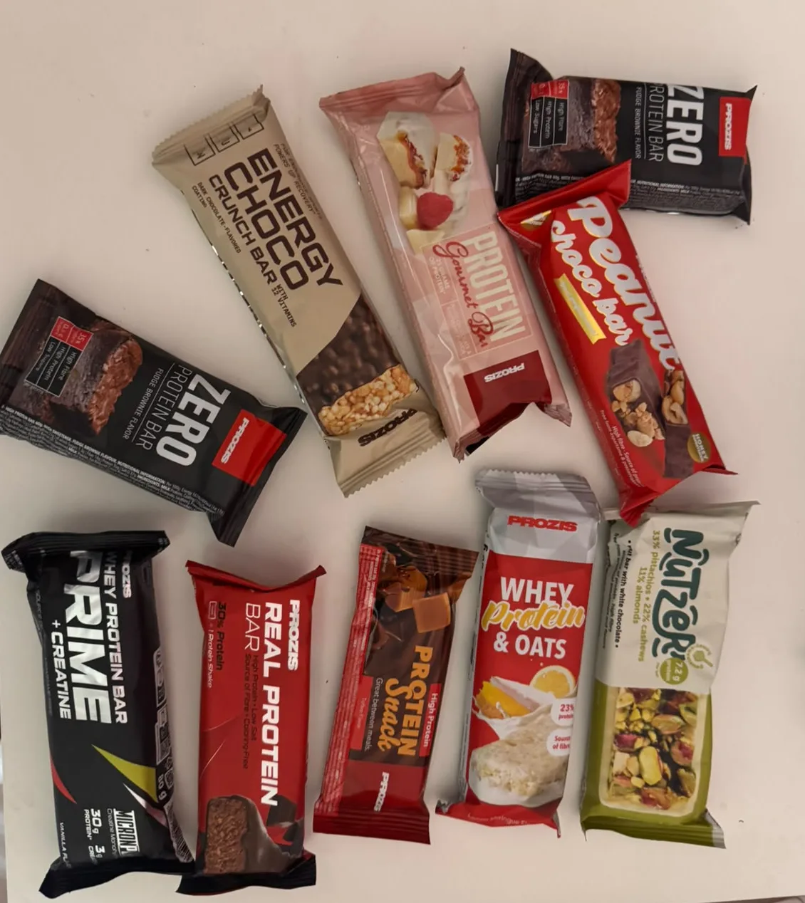 Diez barritas de Prozis cerradas sobre una mesa blanca: dos Zero Protein Bar Fudge Brownie, Energy Choco Crunch Bar, Protein Gourmet Bar, Peanut Choco Bar, Whey Prime Protein Bar + Creatine Vainilla, Real Protein Bar, Protein Snack Toffee, Whey Protein & Oats Lemon Meringue y Nutzer pistacho