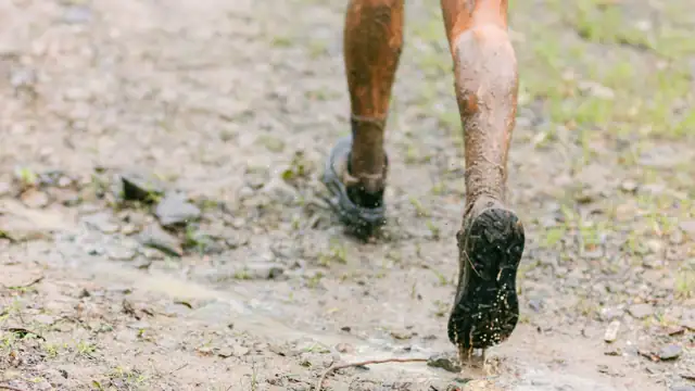 Piernas y zapatillas de trail cubiertas de barro sobre un tramo de grava