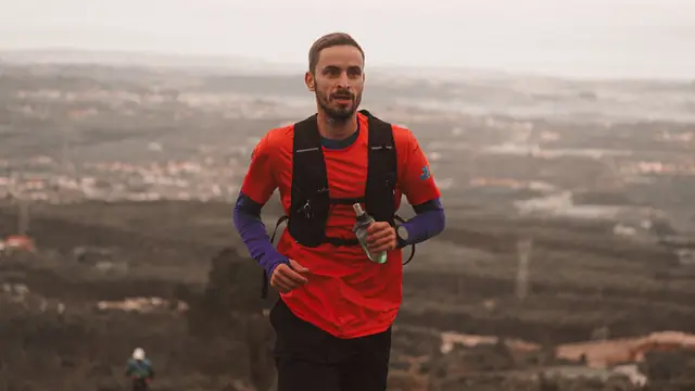 Corredor con chaleco de hidrataci&oacute;n avanzando por un camino de monte con el valle al fondo
